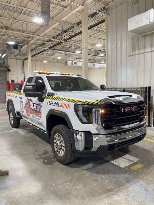 A white GMC pickup truck with 'Junk Busters' branding and hazard stripes parked inside a warehouse.