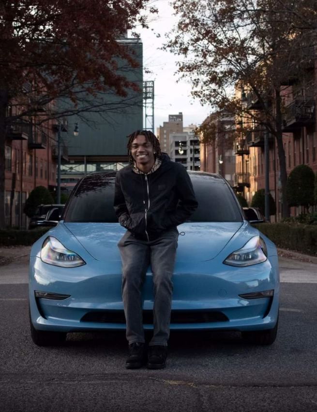 A young man with dreadlocks smiles while sitting on the hood of a light blue Tesla Model 3 on a city street.