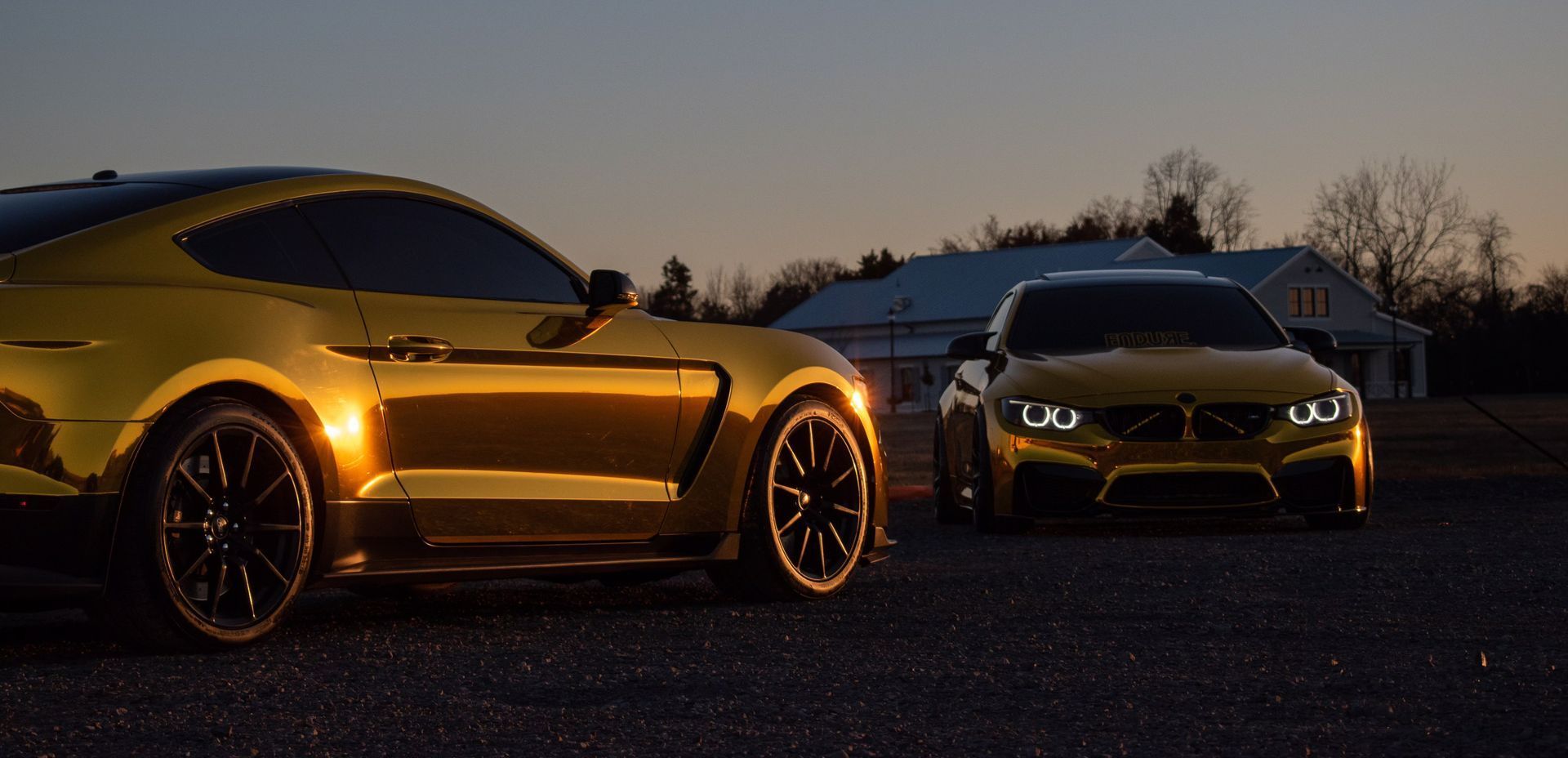 Two gold-painted sports cars are parked side by side on a gravel lot during twilight, their headlights glowing against the fading light.