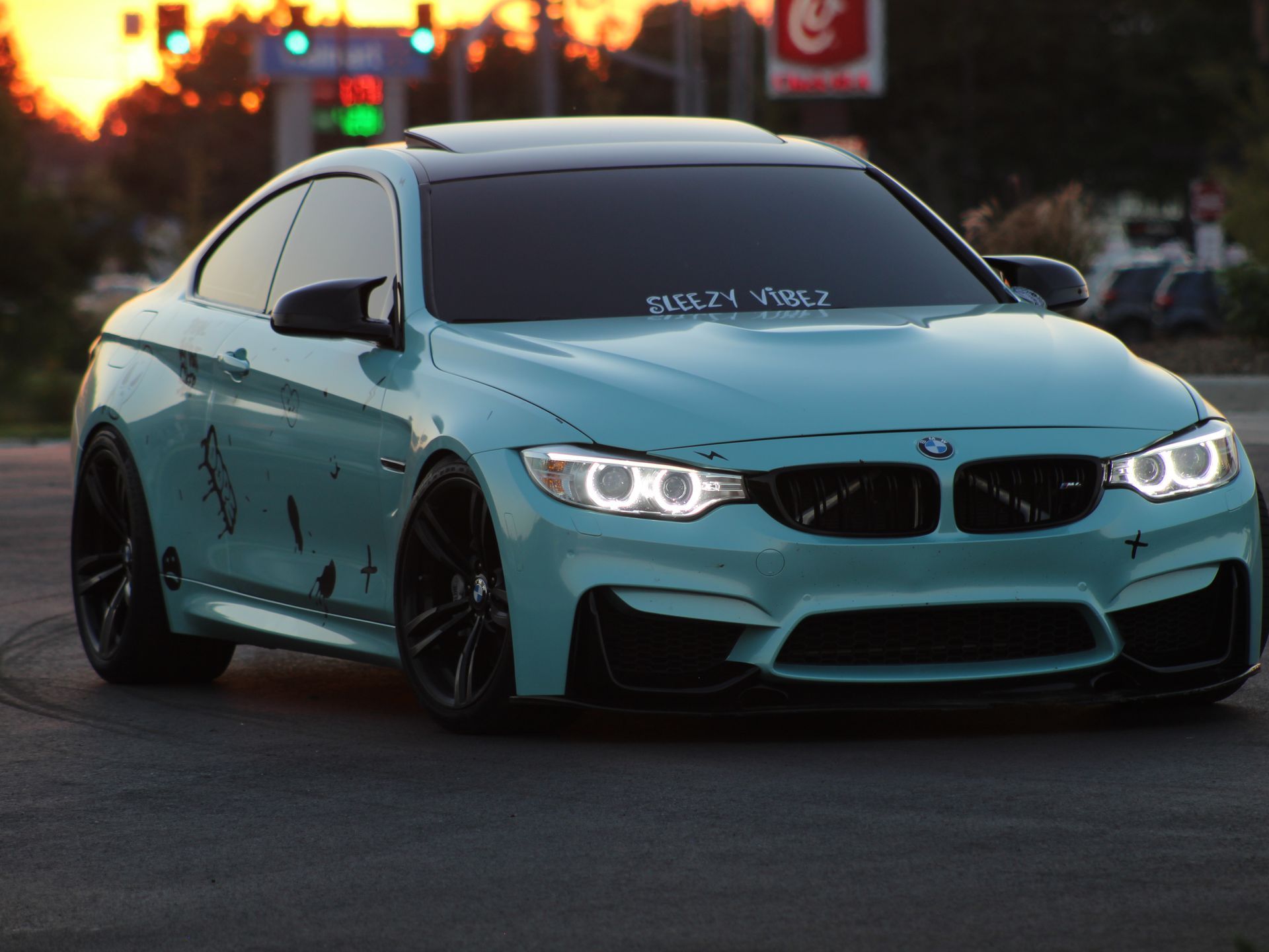 A customized turquoise BMW M4 coupe with black accents and graffiti-style decals, parked at sunset with glowing headlights.