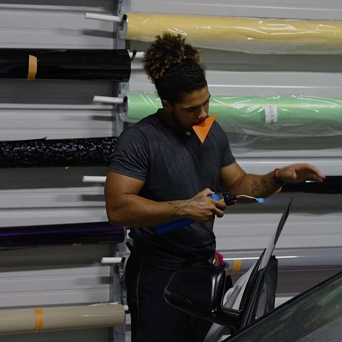 A worker applies tint film to a car window using a heat gun in a workshop.