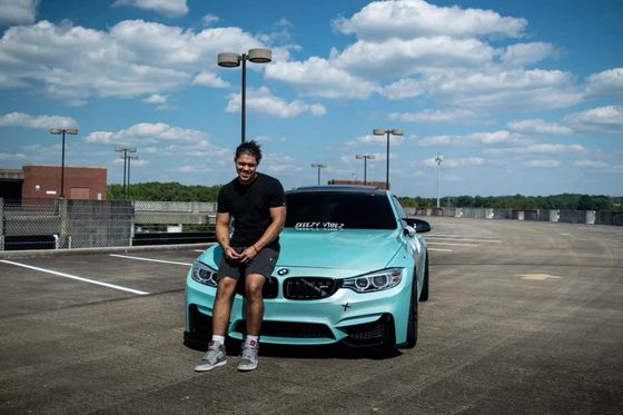 A smiling man leans against a turquoise BMW in an empty parking lot under a partly cloudy sky.