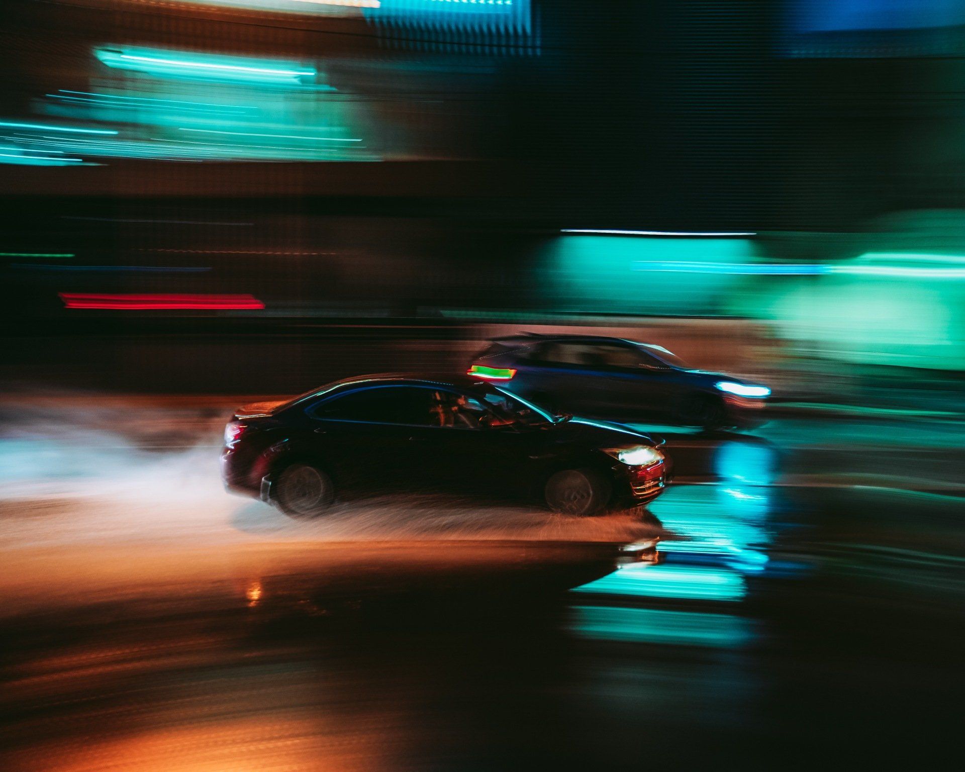 Two cars race through a neon-lit city at night, creating streaks of light and motion blur on wet pavement.