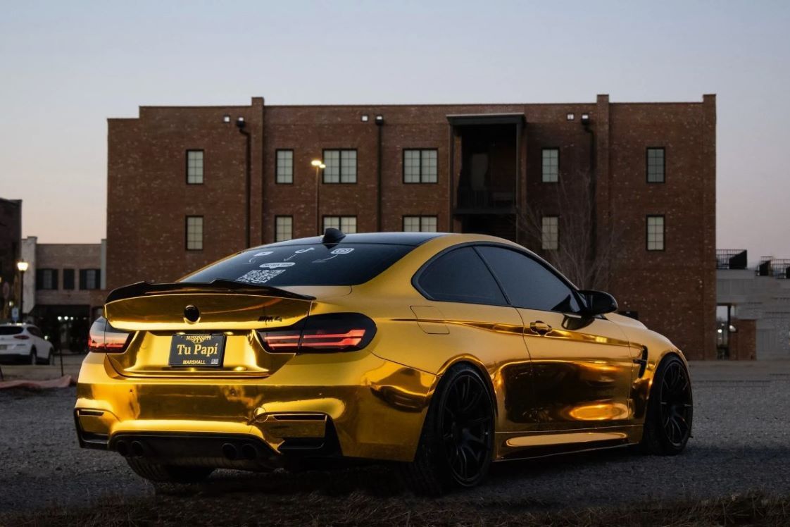 a shiny gold bmw m4 coupe parked in front of a brick building at dusk.