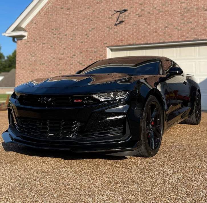 A glossy black Chevrolet Camaro sports car parked on gravel in front of a brick house.